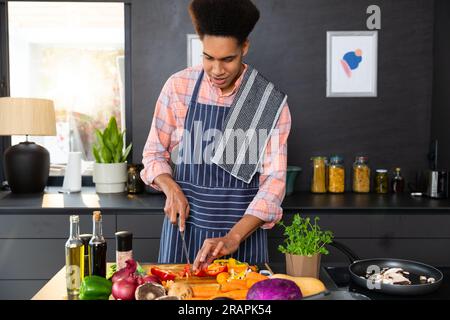 Felice uomo birazziale che indossa il grembiule che prepara il pasto e trita le verdure in una cucina soleggiata. Cibo, cucina, stile di vita sano e vita domestica, inalterati. Foto Stock