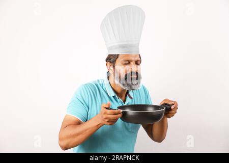 Ritratto di un cuoco indiano barbuto che indossa una t-shirt e un berretto da chef. Espressioni sorridenti su sfondo bianco. Tenere in mano la spatola e il cucchiaio per friggere Foto Stock