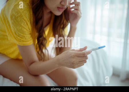 Giovane donna asiatica che sente shock dopo aver guardato il test di gravidanza positivo. Delusa, una bambina incinta non pianificata si siede a letto con la faccia triste e sconvolta Foto Stock