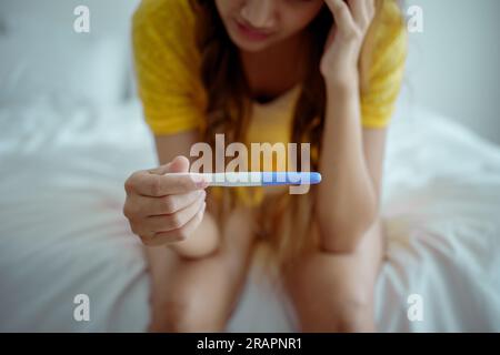 Giovane donna asiatica che sente shock dopo aver guardato il test di gravidanza positivo. Delusa, una bambina incinta non pianificata si siede a letto con la faccia triste e sconvolta Foto Stock