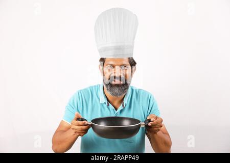 Ritratto di un cuoco indiano barbuto che indossa una t-shirt e un berretto da chef. Espressioni sorridenti su sfondo bianco. Tenere in mano la spatola e il cucchiaio per friggere Foto Stock