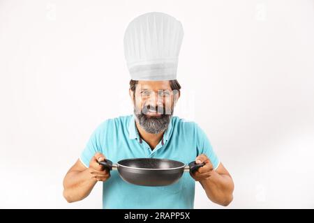 Ritratto di un cuoco indiano barbuto che indossa una t-shirt e un berretto da chef. Espressioni sorridenti su sfondo bianco. Tenere in mano la spatola e il cucchiaio per friggere Foto Stock