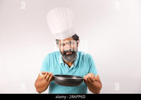 Ritratto di un cuoco indiano barbuto che indossa una t-shirt e un berretto da chef. Espressioni sorridenti su sfondo bianco. Tenere in mano la spatola e il cucchiaio per friggere Foto Stock