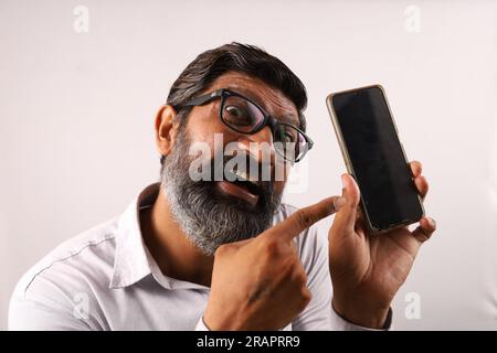 Uomo indiano barbato di mezza età che indossa una camicia che tiene in mano e mostra un telefono cellulare. Espressioni funky su sfondo piano. Foto Stock