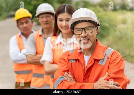 Gruppo di 3 uomini felici e 1 ingegnere donna che indossa tuta arancione, elmetto e giubbotto di sicurezza sorridenti e in piedi all'esterno con le braccia incrociate e la sella Foto Stock