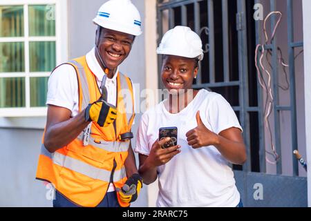 Tecnici maschi e femmine che indossano un casco di sicurezza che reggono i pollici verso l'alto mentre utilizzano uno smartphone, tecnologia wireless Foto Stock