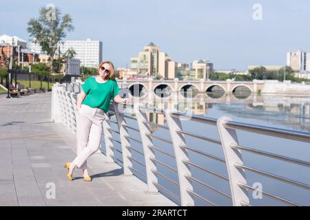 Un ritratto a figura intera di una ragazza felice in una camicetta verde, pantaloni e occhiali da sole che camminano lungo il lungomare. Una ragazza cammina per le strade del Foto Stock