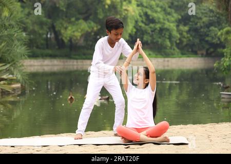 Bambini che fanno esercizi di yoga la mattina presto in un parco pubblico verde. Possiamo vedere il corpo d'acqua. I bambini sono attenti alla salute. Giornata internazionale dello yoga. Foto Stock