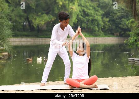 Bambini che fanno esercizi di yoga la mattina presto in un parco pubblico verde. Possiamo vedere il corpo d'acqua. I bambini sono attenti alla salute. Giornata internazionale dello yoga. Foto Stock