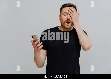Uomo spaventato con t-shirt nera copre mezza faccia, stupito dallo smartphone. Video orribile in mostra, leggendo post sui social media. Isolato su sfondo blu. Foto Stock