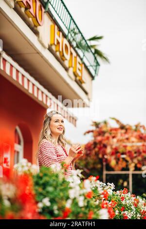 Ritratto in stile retrò di una bella donna con i capelli biondi, in piedi su una terrazza, che tiene una tazza di tè e indossa un blazer a quadri rosso e bianco Foto Stock