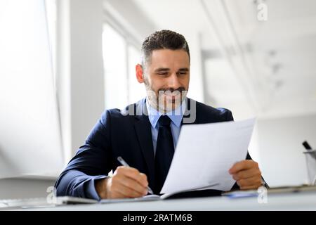 Uomo d'affari positivo di mezza età che lavora con i documenti in ufficio, legge i rapporti finanziari e prende appunti Foto Stock