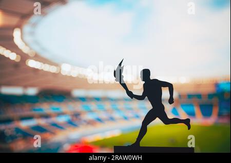 Accendere lo spirito: Silhouette di atleta maschile che trasporta il relè della torcia. Moderno stadio di atletica leggera e campo da golf come sfondo suggestivo. Catturare Essen Foto Stock