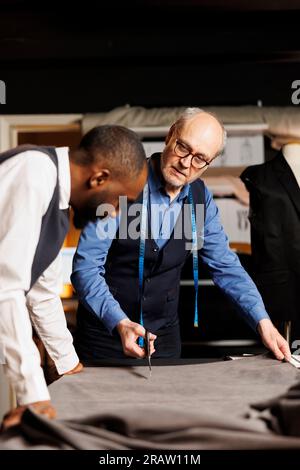 Esperto maestro sartoriale anziano e apprendista sartoriale che taglia i fogli di tessuto per la prossima collezione di abbigliamento su misura. I sartoriali realizzano capi sartoriali eleganti con una lavorazione artigianale raffinata e precisa Foto Stock