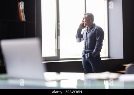 Un uomo d'affari maturo sorridente che parla al telefono cellulare utilizzando un computer portatile in un ufficio moderno. Uomo d'affari di mezza età che chiama da cellulare e guarda un computer che ha una truffa Foto Stock