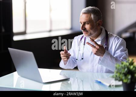 Dottore seduto alla scrivania al suo posto di lavoro e sorridente alla macchina fotografica. Servizio medico perfetto in clinica. Felice futuro della medicina e della sanità. Foto Stock