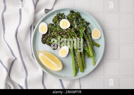 Gustosi broccolini cotti con formaggio, uova di quaglia e limone su un tavolo piastrellato bianco, vista dall'alto Foto Stock