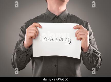 Scuse. Uomo che tiene la carta con la frase mi dispiace su sfondo grigio, primo piano Foto Stock