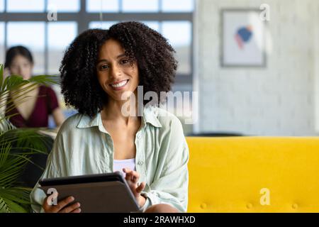 Ritratto di una donna d'affari birazziale e informale che usa un tablet seduto sul divano in ufficio, nello spazio per le copie. Lavoro, affari, comunicazione e ufficio informale, u Foto Stock