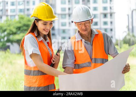 Ingegneri seri maschi e felici con elmetto giallo e bianco che indossano un gilet di sicurezza arancione e osservano il modello di costruzione che stanno bene Foto Stock