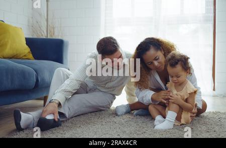 Il calore dei genitori abbracciando e stuzzicando la bambina mentre trascorrono del tempo insieme sul pavimento nel soggiorno di casa. Mamma e papa' usano il tempo libero con b Foto Stock