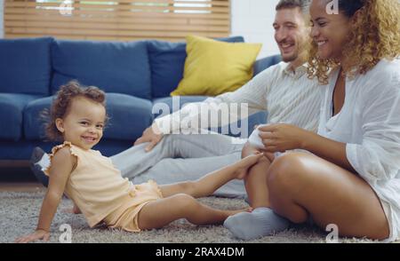 Il calore dei genitori abbracciando e stuzzicando la bambina mentre trascorrono del tempo insieme sul pavimento nel soggiorno di casa. Mamma e papa' usano il tempo libero con b Foto Stock