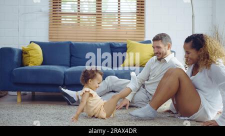 Il calore dei genitori abbracciando e stuzzicando la bambina mentre trascorrono del tempo insieme sul pavimento nel soggiorno di casa. Mamma e papa' usano il tempo libero con b Foto Stock