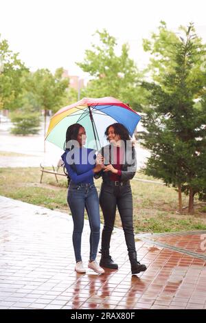 Due donne, afroamericane e latine, che passeggiano in un parco in un giorno di pioggia sotto un ombrello multicolore. Foto Stock