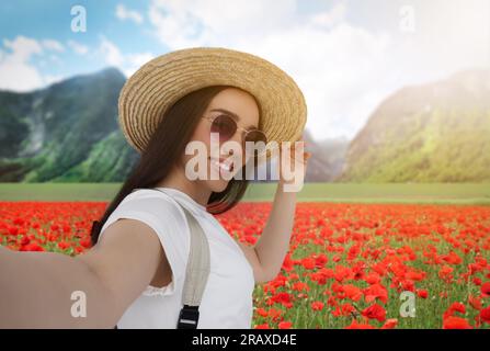 Bella donna in cappello di paglia con occhiali da sole che scatta selfie in fiori di papavero vicino alla montagna Foto Stock