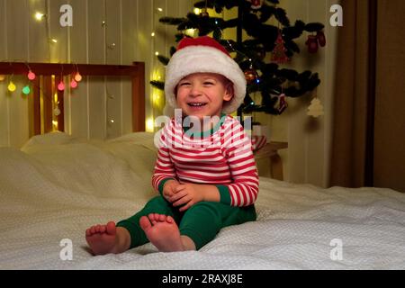 Il ragazzo di Natale a Babbo Natale sorride la sera di casa. Capodanno e festività. Felicità di Natale. Il ragazzo con il cappello di Babbo Natale si diverte, si diverte Foto Stock