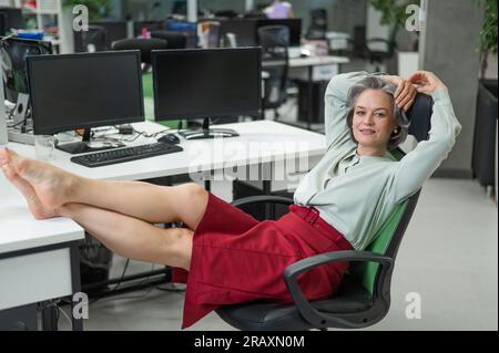 Donna caucasica matura tolse le scarpe e mise i piedi sul desktop in ufficio. Foto Stock