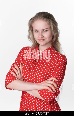 Ritratto di una giovane donna che sorride con le braccia incrociate sul petto guardando la fotocamera. Affascinante modello femminile europeo di 21 anni vestita in abito rosso Foto Stock