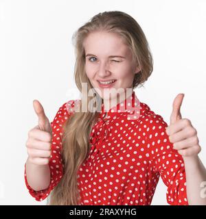 Una giovane donna bionda e sorridente ha scherzosamente chiuso un occhio, mostra i pollici in alto con entrambe le mani. Donna caucasica di 21 anni vestita in abito rosso estivo Foto Stock