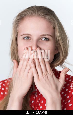 La donna bionda si copre la bocca con le mani e guarda la macchina fotografica. Ritratto di un'autentica modella femminile di 21 anni vestita in abito rosso estivo Foto Stock