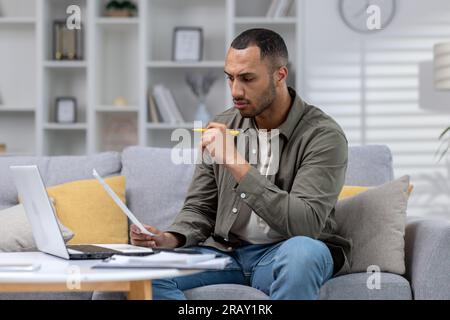 Preoccupato e serio giovane afroamericano che lavora a casa con documenti e bollette. Si siede avvolto sul divano di fronte al portatile e tiene i documenti in mano. Foto Stock