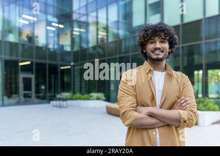 Ritratto di uno studente indiano giovane sicuro di sé, bello e intelligente che si trova all'esterno del campus con le braccia incrociate sul petto e sorridente davanti alla fotocamera. Foto Stock