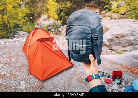 Borsa a compressione piccola e leggera con sacco a pelo in piuma sullo sfondo di una tenda in campeggio e varie attrezzature e oggetti da campeggio Foto Stock