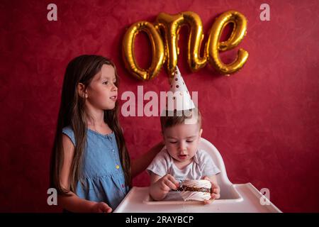 La sorella maggiore si congratula con il fratello minore per il suo primo compleanno. La ragazza bacia gli abbracci da bambino. I bambini si divertono su sfondo rosso con la lamina B. Foto Stock