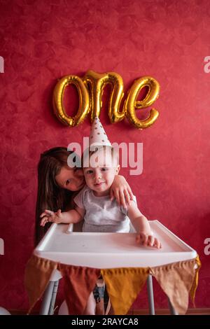 La sorella maggiore si congratula con il fratello minore per il suo primo compleanno. La ragazza bacia gli abbracci da bambino. I bambini si divertono su sfondo rosso con la lamina B. Foto Stock