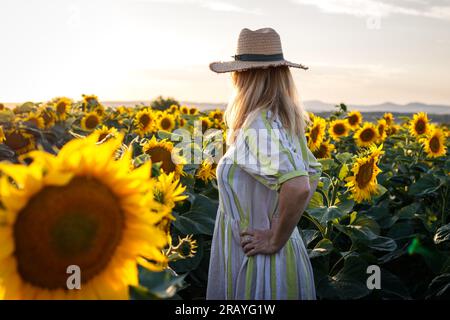 Donna che indossa un abito da sole e un cappello di paglia e si trova in piedi in un campo di girasole. Orgogliosa contadina nella sua fattoria biologica Foto Stock