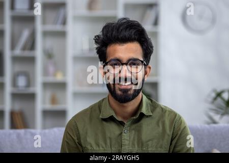 Ritratto ravvicinato di un giovane uomo ispanico sorridente che sorride e guarda la macchina fotografica mentre si siede sul divano nel soggiorno. Foto Stock