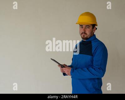 Ritratto del lavoratore o dell'ingegnere in abiti da lavoro blu e casco di sicurezza giallo con spazio per il testo. Un uomo in un cantiere tiene un tablet nella sua h Foto Stock
