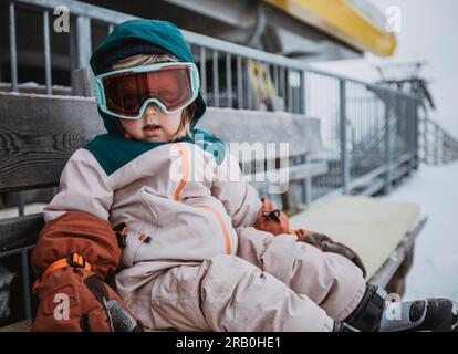 Una bambina in tuta da sci con occhiali da sci siede su una panchina Foto Stock