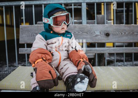 Una bambina in tuta da sci con occhiali da sci siede su una panchina Foto Stock