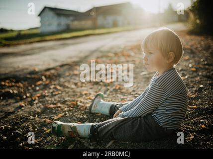 Ragazzo con stivali di gomma si siede a terra di fronte alla casa Foto Stock