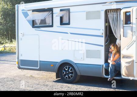 Concetto di vacanza di viaggio e stile di vita in camper. Una giovane donna di mezza età seduta sulla porta del suo camper di casa alternativo camper camper che si gode il parcheggio a destinazione. Vanlife gente fuori rete Foto Stock