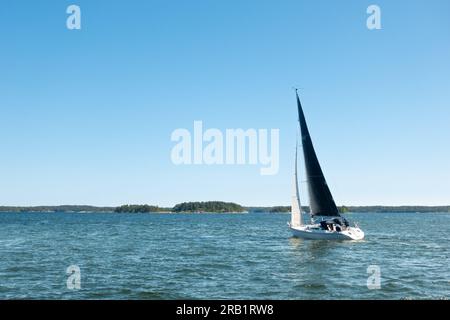 Barca a vela con vela nera vista dalla poppa che naviga sulle acque turchesi in una giornata di cielo azzurro nell'arcipelago di Stoccolma in Svezia Foto Stock