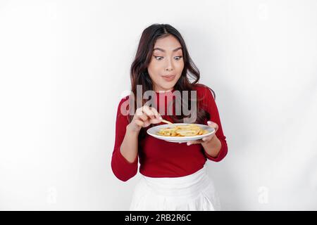 Giovane donna affamata che indossa abiti informali, tiene un piatto con fast food, patatine fritte isolate su sfondo bianco, ritratto in studio. Lo stile di vita delle persone Foto Stock