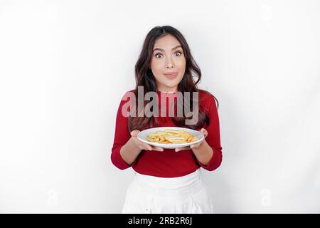 Giovane donna affamata che indossa abiti informali, tiene un piatto con fast food, patatine fritte isolate su sfondo bianco, ritratto in studio. Lo stile di vita delle persone Foto Stock