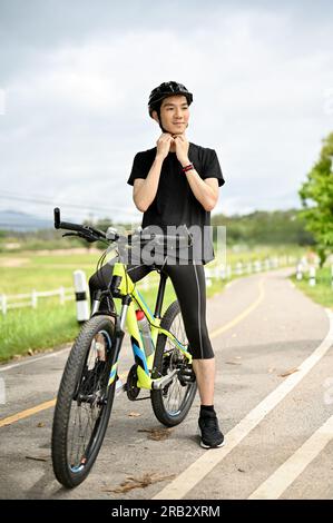 Un ritratto di un giovane asiatico felice e affascinante che indossa abbigliamento sportivo togliendosi il casco da bici dopo aver messo in bicicletta il casco prima di andare in bicicletta Foto Stock